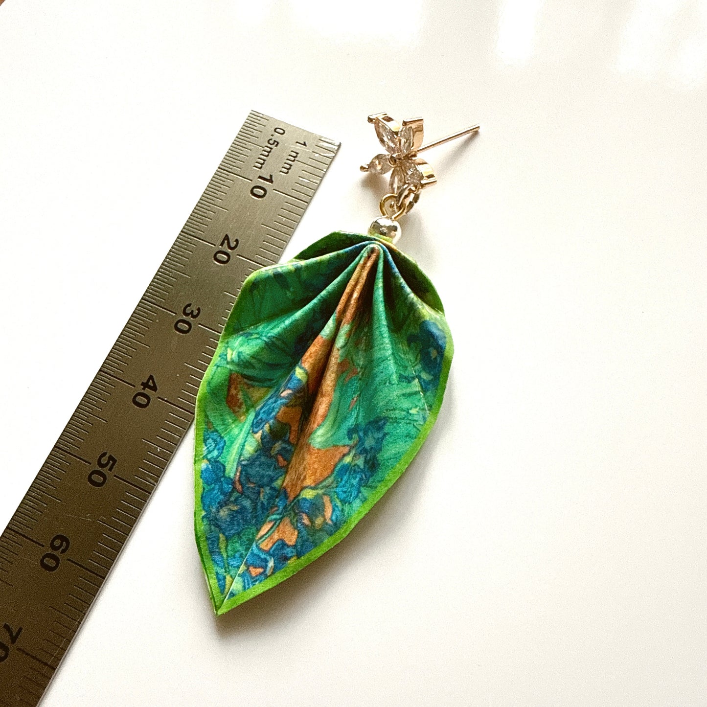 Leaf-shaped pendant with floral design next to a ruler on a white background