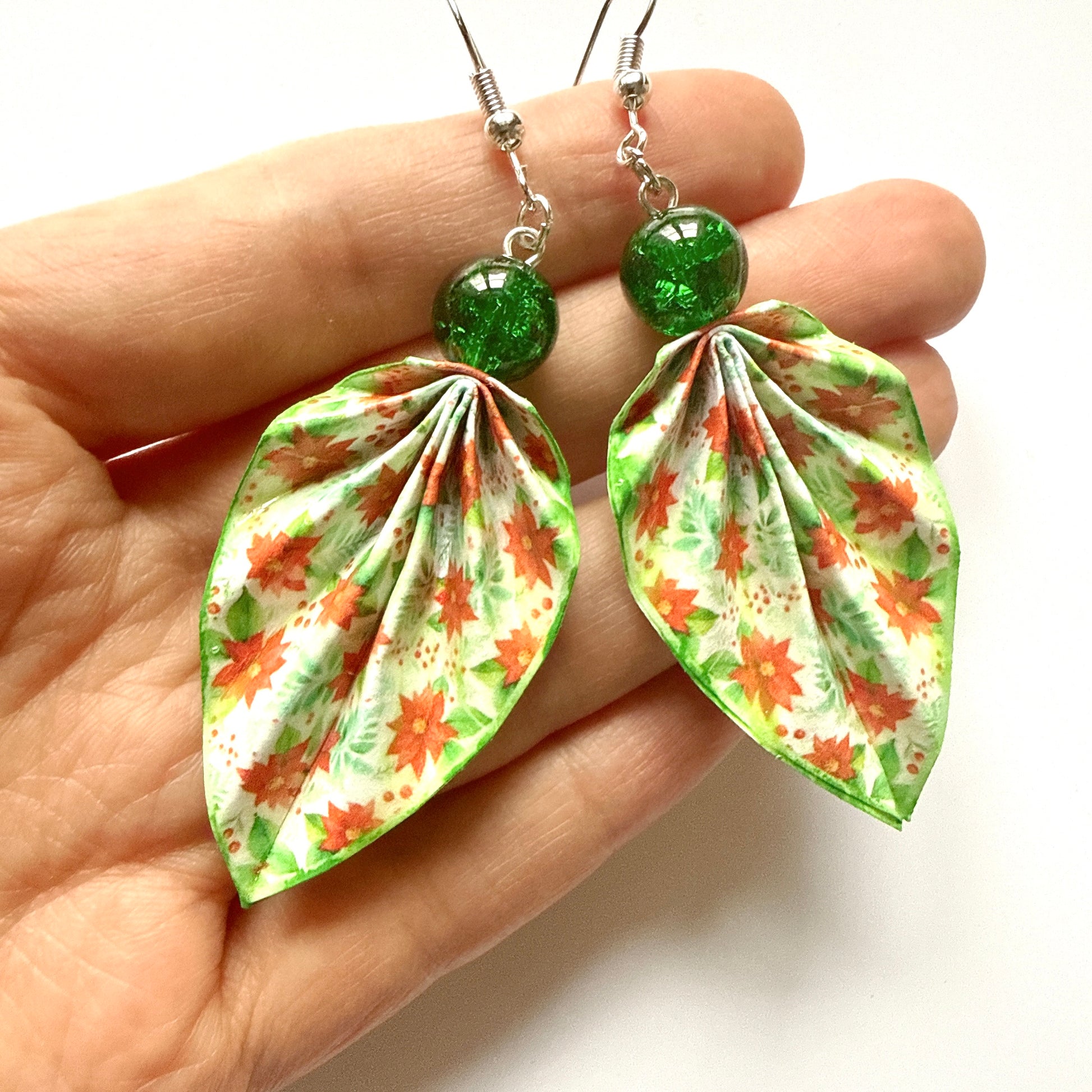 Floral leaf-shaped earrings with green beads held in a hand against a white background